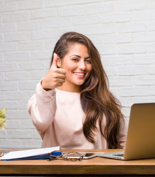 Portrait of young student latin woman sitting on her desk cheerful and excited, smiling and raising her thumb up, concept of success and approval, ok gesture